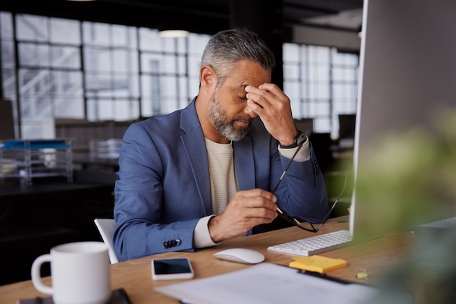 Mature businessman holding his head in stress, sitting at a desk with computer and documents. Indian manager working late and worried for company deadline. 